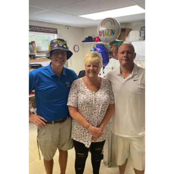 Three smiling people, two men and one woman, stand together in what appears to be a deli.