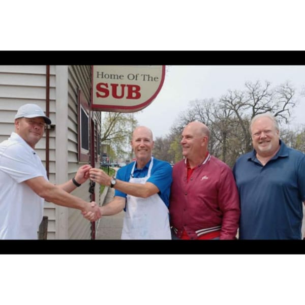 Four men outside Lee's Deli, two shaking hands, with a "Home Of The SUB" sign visible.