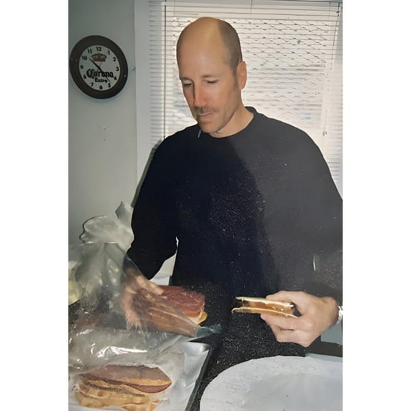 A man preparing a sub sandwich, holding a piece of bread and meat, with a Corona clock in the background.