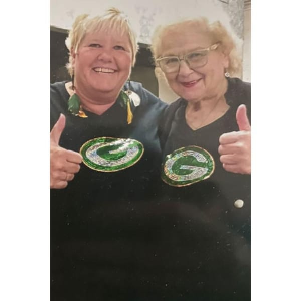 Two smiling women in black shirts with green and silver G logos give thumbs up.