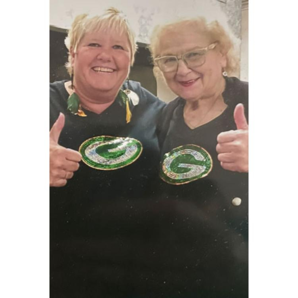 Two smiling women in black shirts with green and silver G logos give thumbs up.